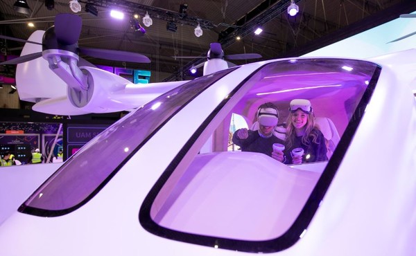 MWC SKT 전시관에서 현지 모델들이 UAM 모형에 탑승해 2030년의 서울을 배경으로 비행하는 서비스를 체험하고 있는 모습. 사진출처 = SK텔레콤